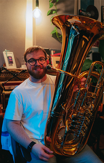 Ein junger Mann mit Brille und Bart sitzt in einem gemütlich beleuchteten Raum und hält eine glänzende goldene Tuba. Er trägt ein weißes T-Shirt und lächelt in die Kamera. Im Hintergrund sind ein aufgeschlossenes Klavier, Pflanzen und Schallplatten zu sehen. Es handelt sich um den Tubisten Frederik Bauersfeld.