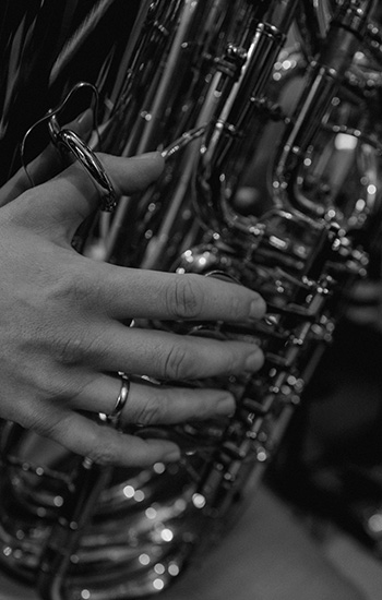 Nahaufnahme einer linken Hand mit Ring, die auf den Ventilen einer glänzenden Tuba liegt. Das Bild ist in Schwarz-Weiß gehalten und zeigt feinste Details der Fingerhaltung und der komplexen Ventilmechanik des Instruments.
