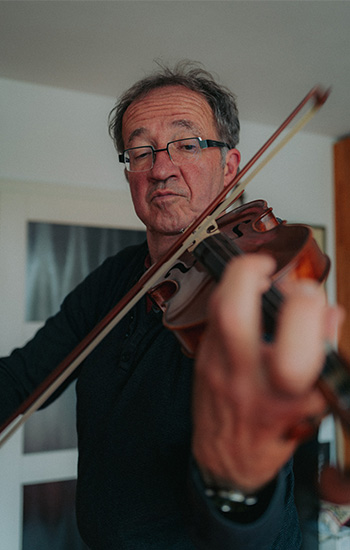 Ein älterer Mann, Ulrich Menke, spielt konzentriert Geige in einem Innenraum. Er trägt eine dunkle Brille und ein dunkles Oberteil. Die Kamera ist nah an seiner linken Hand positioniert, wodurch der Fokus auf den Saiten und dem Griffbrett der Geige liegt. Der Hintergrund zeigt eine helle Wand und ein unscharfes Bild oder Fenster.