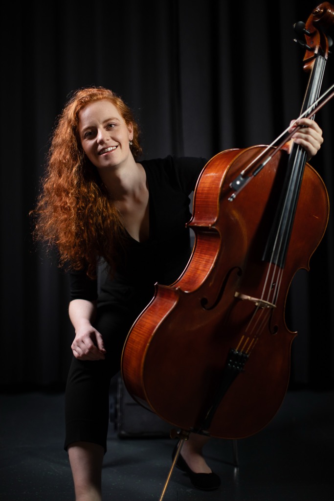 Teresa Wenhart sitzt neben ihrem Cello und vor einem Schwarzen Hintergrund.